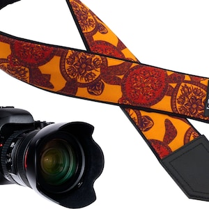 May include: A camera strap with a colorful orange and red patterned design featuring a repeating mandala pattern. The strap is made of fabric and has black leather ends. The text "InTePro" is printed on one of the ends.