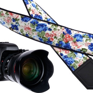 Può includere: Un cinturino per fotocamera con sfondo bianco e un motivo floreale con fiori blu e rosa. Il cinturino è realizzato in tessuto nero con una finitura simile alla pelle.