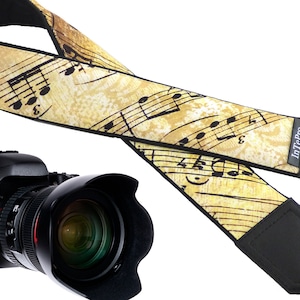 Può includere: Una tracolla per fotocamera con un motivo a note musicali su sfondo giallo. La tracolla è realizzata in tessuto nero con logo InTePro nero. La tracolla è fissata a una fotocamera reflex digitale nera con un obiettivo.