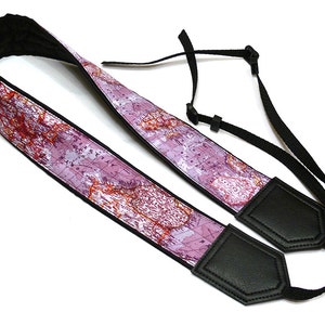 May include: A pink and white camera strap with a world map design. The strap has black leather ends and adjustable black straps.
