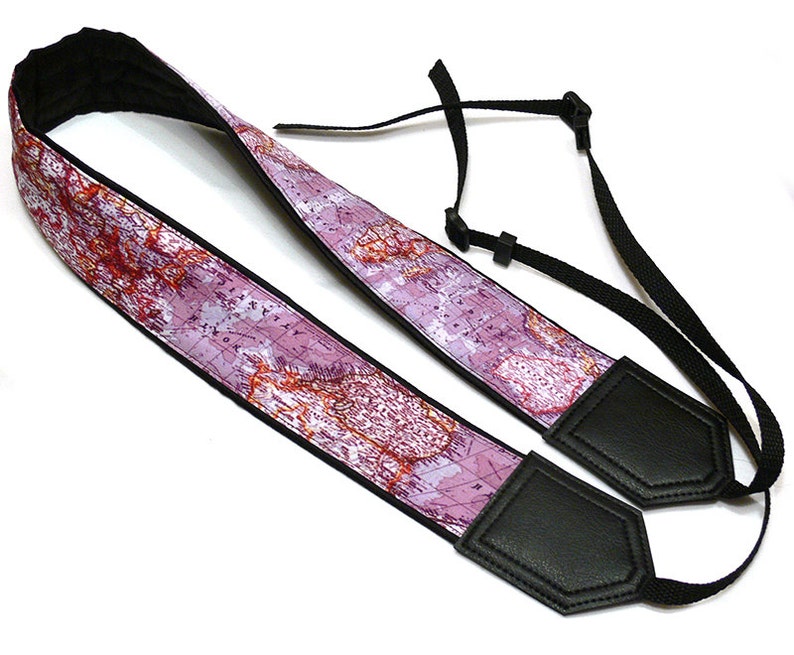 May include: A pink and white camera strap with a world map design. The strap has black leather ends and adjustable black straps.