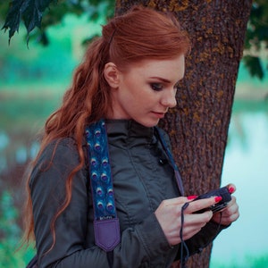 May include: A woman with long, red hair is standing near a tree. She is wearing a dark blue jacket with a peacock-patterned camera strap. She is holding a small black camera in her hands.