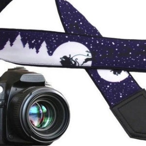 May include: A black camera with a large lens and a blue and white camera strap featuring a winter scene design. The strap has a black leather pad.