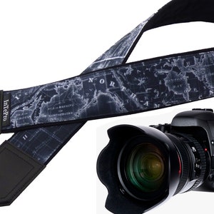 May include: A black and white camera strap with a map design. The strap is made of a soft, padded material and has a leather-like finish. The strap is adjustable and has a quick-release buckle. The strap is shown with a black camera with a large lens.