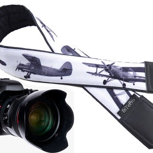 Può includere: Un cinturino per fotocamera bianco e nero con un design stampato di aerei. Il cinturino ha dettagli in pelle nera e il testo "InTePro" stampato sul cinturino.