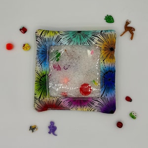 Könnte beinhalten: Ein sensorisches Kissen mit einem transparenten Fenster, gefüllt mit weißen Perlen und kleinen, bunten Formen. Das Kissen hat einen lebendigen, mehrfarbigen Blumenmuster-Rand. Kleine, dekorative Verzierungen in Tier- und Lebensmittelform sind verstreut.