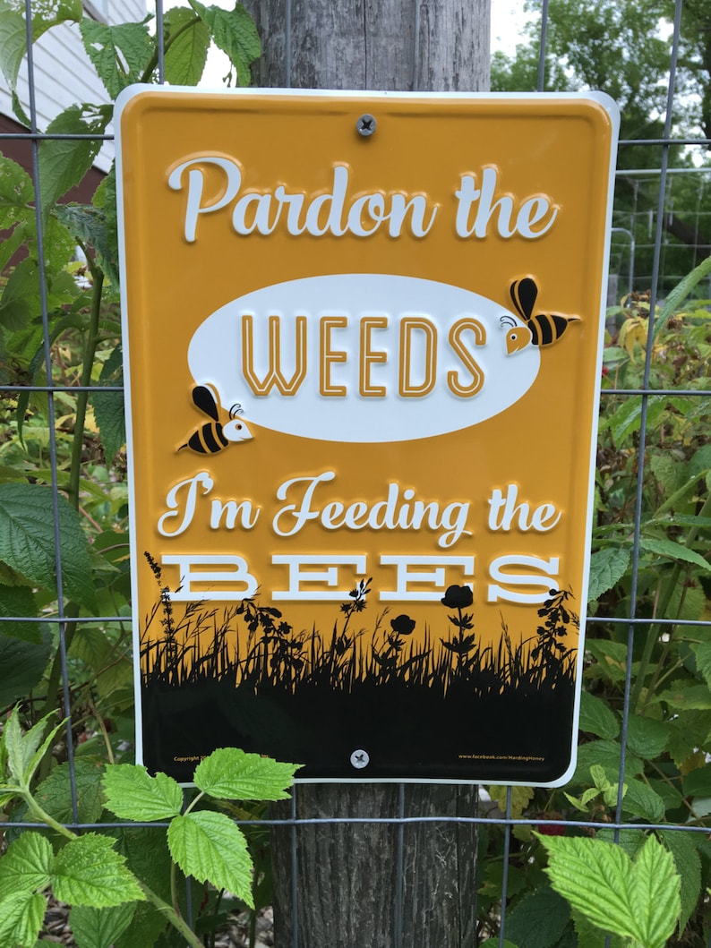 以下が含まれることがあります： 「Pardon the weeds I'm feeding the bees」という文字と、漫画の蜂のイラストが描かれた黄色の金属製の看板。看板は木製のフェンスに取り付けられています。