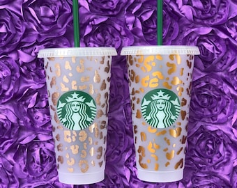 Könnte beinhalten: Zwei durchsichtige Plastikbecher mit Deckel und Strohhalmen. Die Becher sind mit einem pinkfarbenen und goldenen Leopardenmuster und dem Starbucks-Logo verziert.