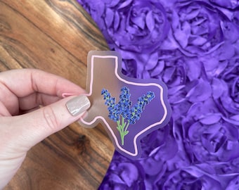 May include: A clear sticker of the state of Texas with a purple flower bouquet inside. The sticker is on a purple background.