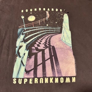 May include: A black t-shirt with a graphic print of a walkway with a checkered pattern leading up to a building with a starry night sky in the background. The text "SOUNDGARDEN" is at the top of the graphic and the text "SUPERUNKNOWN" is at the bottom of the graphic.