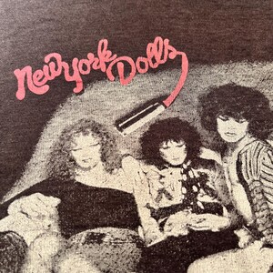 Puede incluir: Una camiseta marrón con una imagen en blanco y negro de la banda "New York Dolls" impresa en la parte delantera. Los miembros de la banda aparecen sentados en un sofá.