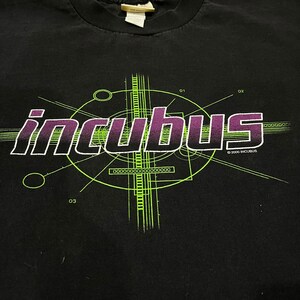 Puede incluir: Camiseta negra con un gráfico verde y morado de la banda Incubus. El gráfico presenta el nombre de la banda en una fuente estilizada con formas geométricas y líneas. El texto "© 2000 INCUBUS" está impreso en blanco en la parte inferior del gráfico.