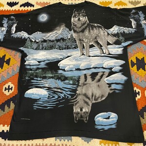 K&ouml;nnte beinhalten: Schwarzes T-Shirt mit einem Wolf, der in einer verschneiten Waldszene zum Mond heult. Die Reflexion des Wolfes ist im Wasser darunter zu sehen.