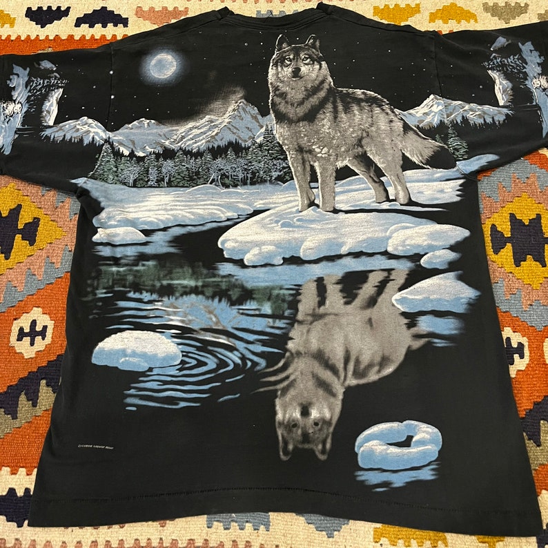 K&ouml;nnte beinhalten: Schwarzes T-Shirt mit einem Wolf, der in einer verschneiten Waldszene zum Mond heult. Die Reflexion des Wolfes ist im Wasser darunter zu sehen.