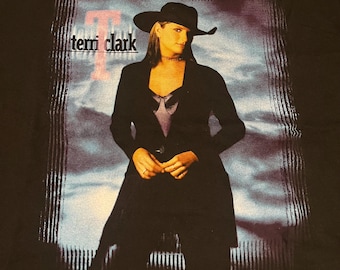 May include: Black t-shirt featuring a portrait of Terri Clark. The image shows a woman in a black hat and blazer, with the name "terri clark" printed in white and a pink "T" in the background. The background has a blue and purple gradient.