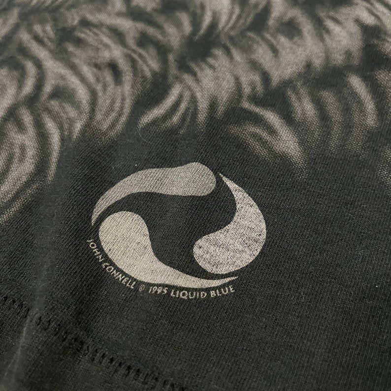 K&ouml;nnte beinhalten: Ein schwarzes T-Shirt mit einem wei&szlig;en Wirbelmuster und einem wei&szlig;en Logo mit der Aufschrift "John Connell &copy; 1995 Liquid Blue".
