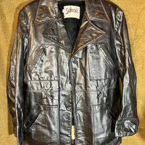 May include: Black leather jacket with a notched collar, four front pockets, and decorative stitching. The jacket has a button closure and a label that reads "Schott". The jacket is hanging on a wooden hanger.
