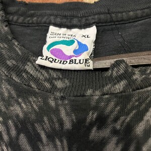 K&ouml;nnte beinhalten: Schwarzes T-Shirt mit einem Batik-Muster und einem Liquid Blue-Etikett. Das Etikett ist wei&szlig; mit blauen und violetten Akzenten und tr&auml;gt die Aufschrift "Liquid Blue" in wei&szlig;en Buchstaben. Das Etikett tr&auml;gt au&szlig;erdem die Aufschrift "Made in USA" und "Care on Reverse" in kleineren wei&szlig;en Buchstaben. Das Etikett hat ein Gr&ouml;&szlig;enetikett mit der Aufschrift "XL".