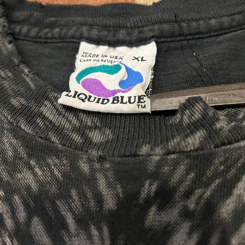 K&ouml;nnte beinhalten: Schwarzes T-Shirt mit einem Batik-Muster und einem Liquid Blue-Etikett. Das Etikett ist wei&szlig; mit blauen und violetten Akzenten und tr&auml;gt die Aufschrift "Liquid Blue" in wei&szlig;en Buchstaben. Das Etikett tr&auml;gt au&szlig;erdem die Aufschrift "Made in USA" und "Care on Reverse" in kleineren wei&szlig;en Buchstaben. Das Etikett hat ein Gr&ouml;&szlig;enetikett mit der Aufschrift "XL".