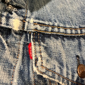 Puede incluir: Primer plano de una chaqueta vaquera azul con una etiqueta roja de Levi's y un botón de cobre. El denim tiene un aspecto descolorido y desgastado.