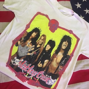以下が含まれることがあります： Motley Crueのバンドの黄色と赤のグラフィックが描かれた白いTシャツ。グラフィックは、頭蓋骨の形をしたフレームの中にバンドメンバーをフィーチャーしています。