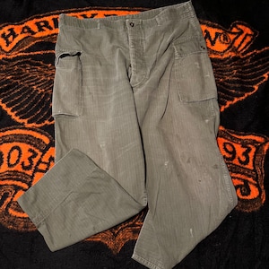 Puede incluir: Pantalones cargo verde oliva con dos bolsillos delanteros y dos laterales. Los pantalones tienen cierre de botón y pernera recta. La tela muestra signos de desgaste, lo que añade a la estética vintage. Miden aproximadamente 100 cm de largo.