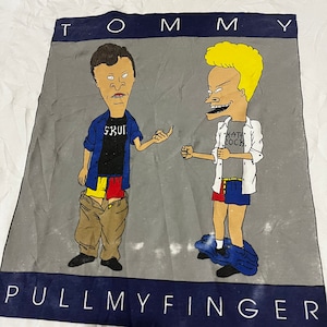 May include: A white t-shirt with a gray background featuring two cartoon characters. The characters are wearing shorts and have the text "TOMMY" at the top and "PULL MY FINGER" at the bottom.