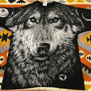 K&ouml;nnte beinhalten: Schwarzes T-Shirt mit einem gro&szlig;en, detaillierten Druck eines Wolfskopfes. Der Wolf hat gr&uuml;ne Augen und eine schwarze Nase. Der Hintergrund des Drucks ist ein dunkler, sternen&uuml;bers&auml;ter Nachthimmel. Das T-Shirt hat ein kleines, kreisf&ouml;rmiges Logo in der unteren rechten Ecke.