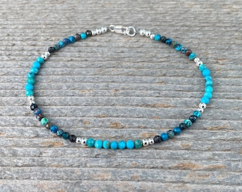 May include: An anklet featuring small, round turquoise and dark blue beads interspersed with silver-colored beads. The anklet has a silver clasp and is displayed on a wooden surface.