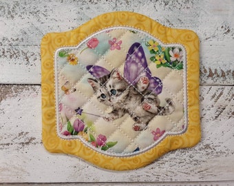 May include: A yellow quilted hot pad with a white border and a whimsical design of a gray kitten with purple butterfly wings holding a star wand. The kitten is surrounded by colorful flowers and butterflies.