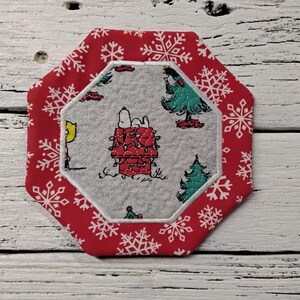 May include: An octagonal Christmas coaster with a red border featuring white snowflakes. The center has a gray background with Snoopy on his doghouse decorated with Christmas lights, and a Christmas tree. A yellow bird is also visible.