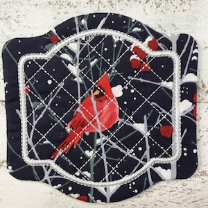 May include: A blue quilted fabric with a white embroidered frame and a red cardinal bird perched on a snowy branch. The fabric has a wintery design with white snowflakes and red berries.
