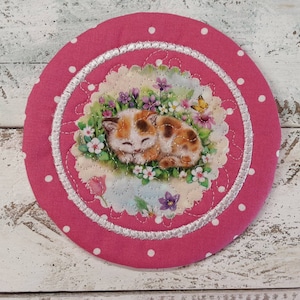 May include: A pink fabric coaster with white polka dots and a white satin trim. The center features a circular design with a sleeping kitten surrounded by flowers.