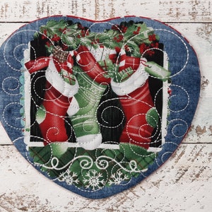 May include: A heart-shaped quilted coaster with a blue denim border and a festive Christmas stocking design. The design features three red and green stockings with white trim and a sprig of holly.
