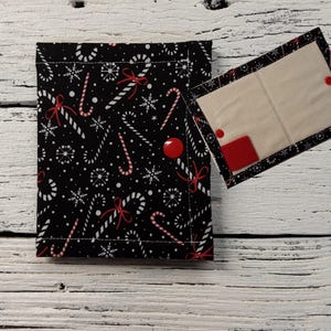 May include: A black fabric wallet with a red and white candy cane pattern. The wallet has a flap that opens to reveal a white fabric interior with two pockets. The wallet is closed with a red button.