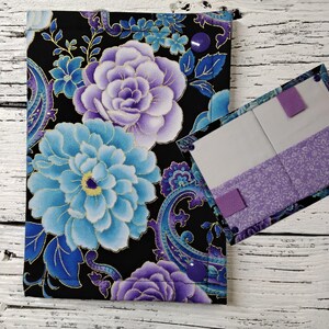 May include: A floral fabric book cover with a matching interior pocket. The cover features large blue and purple flowers on a black background. The interior has a light purple floral pattern and two purple elastic closures.
