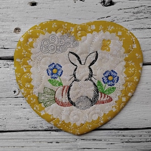 May include: Heart-shaped quilted trivet with a yellow floral border and a white center. The center features an embroidered bunny, two blue flowers, and a carrot. The trivet is on a white wooden surface.
