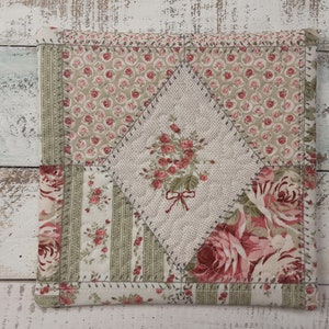 May include: A square patchwork cushion with a diamond-shaped center featuring a floral design. The cushion is made of a variety of fabrics in shades of pink, green, and white.