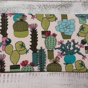 May include: A beige fabric pouch with a colorful cactus print. The pouch has a green, blue, and pink cactus design with a white background. The pouch has two green buttons.
