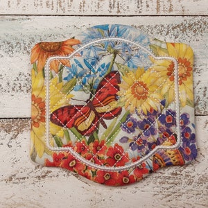 May include: A white fabric coaster with a floral and butterfly design. The coaster has a white stitched border with a scalloped edge. The design features yellow, red, blue, and purple flowers with a large butterfly in the center.