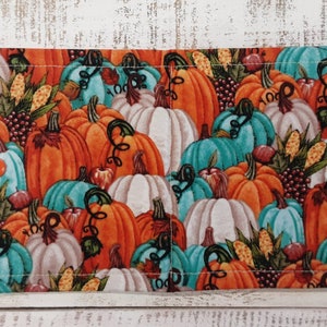 May include: A rectangular fabric item with a vibrant pumpkin pattern. The design features orange, teal, and white pumpkins with green vines, interspersed with corn and small red fruits. Two orange buttons secure the item.
