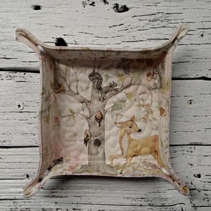May include: A square, fabric catchall tray with a woodland scene. The tray features a deer, squirrels, birds, and a tree on a quilted, light pink background. The tray has curved edges and a light pink trim.