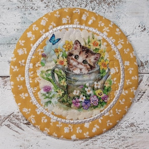 May include: A yellow fabric coaster with a white border and a floral design featuring a kitten inside a watering can. The kitten is looking at the viewer with green eyes.
