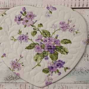 May include: A heart-shaped quilted hot pad with a floral design featuring purple flowers and green leaves on a white background.