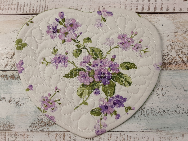 May include: A heart-shaped quilted hot pad with a floral design featuring purple flowers and green leaves on a white background.