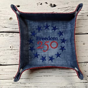 May include: A square fabric catch-all tray with a blue denim interior featuring an embroidered circle of dark blue stars surrounding the text Freedom 250 in red and blue thread. The tray has raised corners secured with white fasteners.