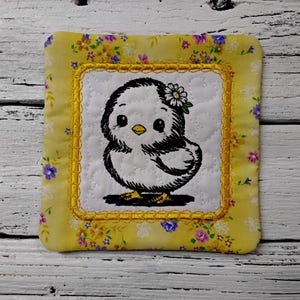 May include: Square yellow fabric coaster with a floral pattern and a white center. The center features a black and white embroidered chick with a flower on its head. The coaster has a gold border.