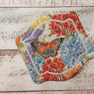 May include: A white fabric pot holder with a floral design featuring a butterfly, red flowers, and blue flowers. The pot holder has a white stitched border.