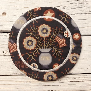 May include: Round fabric coaster with a patriotic design. The coaster features a dark background with floral and star patterns, a vase with the number "1776", and American flags. The edge is trimmed with white stitching.