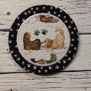 May include: Round quilted coaster with a black border featuring white paw prints. The center has a cream background with a dog and boots illustration. The dog is light brown and the boots are brown and tan.